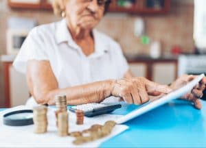 Elderly checking her expenses and looking worried, sitting at a table with bills and a calculator in front of her.